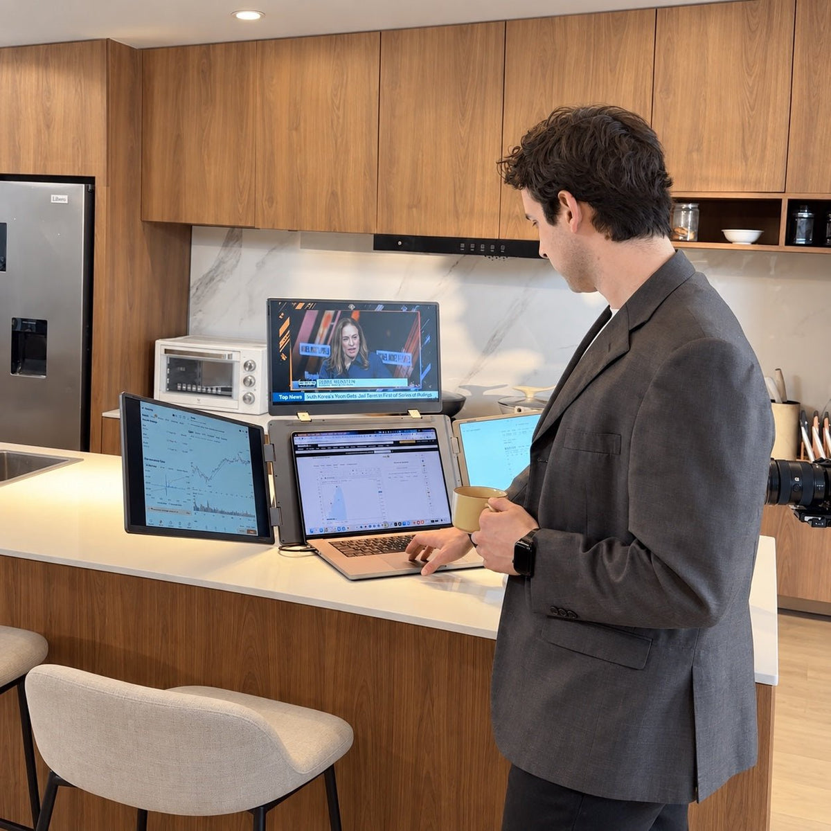 Person holding a mug while using MagHub Quad Max at a kitchen counter, with financial charts and a news video across the laptop and three displays.